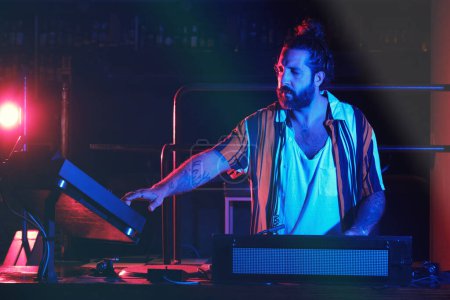 Foto de Varón con barba hábil en ropa casual tocando música mientras está de pie cerca de DJ profesional en un club nocturno moderno con iluminación tenue - Imagen libre de derechos