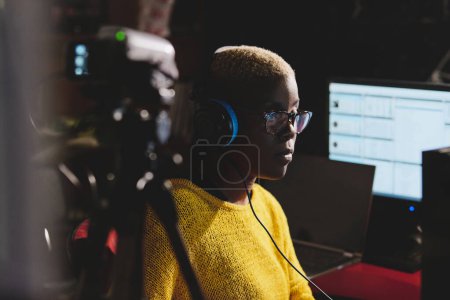 Téléchargez les photos : Vue latérale de dame aux cheveux courts noirs dans des écouteurs et des lunettes regardant loin contre l'ordinateur pendant le travail dans un studio de radiodiffusion - en image libre de droit