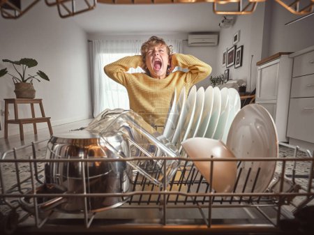 Foto de Muchacho molesto gritando y tocando el cabello mientras está de pie cerca de lavavajillas moderno con varios utensilios de cocina limpia en la cocina ligera en casa - Imagen libre de derechos