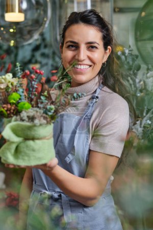 Téléchargez les photos : Femme joyeuse en tablier bleu souriant et regardant la caméra tout en travaillant dans la boutique florale et démontrant bouquet arrangé de fleurs en fleurs - en image libre de droit