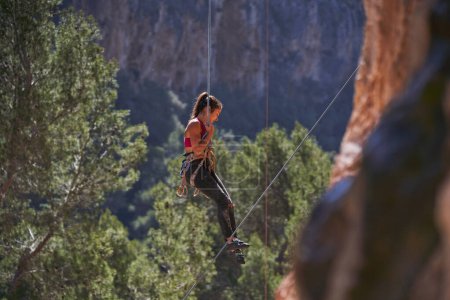Téléchargez les photos : Vue latérale du grimpeur féminin en tenue de sport et harnais de sécurité avec diverses carabines rappelant la falaise sur la corde et regardant vers le bas - en image libre de droit