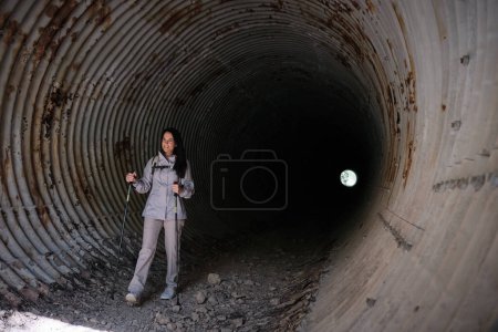 Photo for Full body of active Hispanic female with walking poles standing in long dark tunnel with shabby rough walls during trip - Royalty Free Image
