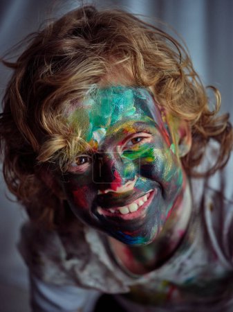 Photo for From above of happy boy with curly hair and colorful painted face looking away with smile while sitting in light room - Royalty Free Image