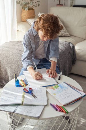 Foto de Colegial preadolescente enfocado en ropa casual sentado cerca del escritorio con cuadernos y dibujo con lápiz en papel en casa - Imagen libre de derechos