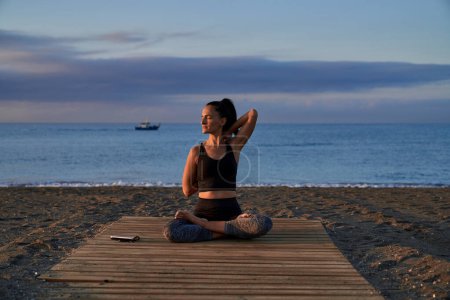 Photo for Full body of peaceful Hispanic female with closed eyes doing Gomukhasana posture while sitting on shore near sea during yoga training - Royalty Free Image