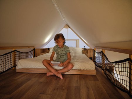 Photo for Full body of barefoot child with legs crossed in bedroom holding notebook sitting on comfortable bed under roof of glamping tent - Royalty Free Image