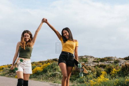 Foto de Felices novias multiétnicas cogidas de la mano mientras caminan por la carretera con longboards - Imagen libre de derechos