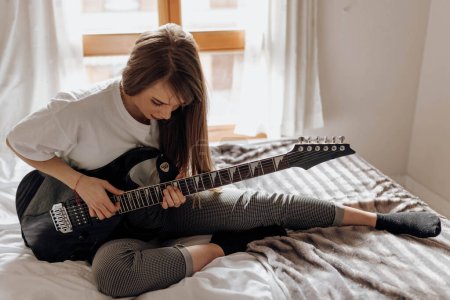 Téléchargez les photos : Vue latérale de Jeune femme brune jouant de la guitare sur le lit - en image libre de droit