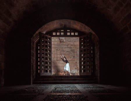 Photo for Full body of anonymous female ballet dancer in white tutu and bodysuit dancing in dark passage of aged building near brick wall and gates - Royalty Free Image