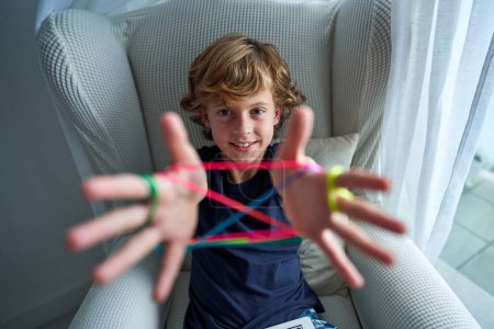Téléchargez les photos : Garçon heureux regardant la caméra avec le sourire tout en jouant jeu de corde coloré avec les bras tendus dans un fauteuil confortable à la maison - en image libre de droit