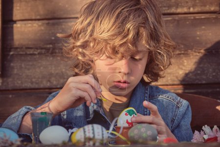 Téléchargez les photos : Petit garçon peignant des œufs pour Pâques à table sur fond flou le jour d'été ensoleillé - en image libre de droit