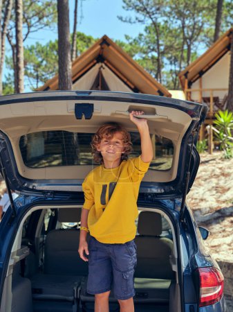 Téléchargez les photos : Enfant souriant tenant la porte du coffre de la voiture près du camping glamping dans la nature pendant le voyage d'été par une journée ensoleillée - en image libre de droit