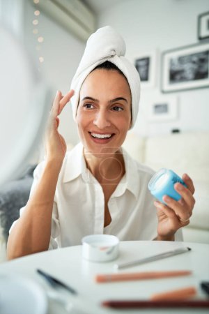 Téléchargez les photos : Femme adulte joyeuse avec serviette sur la tête appliquant crème hydratante pour le visage tout en faisant la procédure de beauté du matin à la maison devant le miroir - en image libre de droit