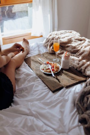 Foto de Mujer joven comiendo gofres apetitosos y bebidas sabrosas en bandeja en la cama - Imagen libre de derechos