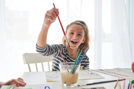 Téléchargez les photos : Fille souriante montrant pinceau coloré regardant la caméra tout en peignant à la table blanche avec un verre d'eau dans la pièce lumineuse - en image libre de droit