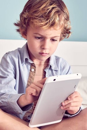Photo for Crop attentive curly haired boy sitting on bed and using modern tablet in morning - Royalty Free Image