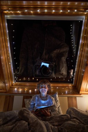 Foto de Contenido niño jugando juego en la tableta y mirando a la cámara mientras está acostado en la cama cómoda cerca de la ventana en la noche - Imagen libre de derechos