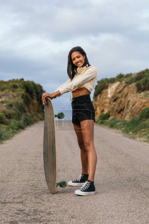 Photo for Full body side view of young smiling ethnic cheerful female standing with longboard and headphones on neck looking at camera between hills under cloudy sky - Royalty Free Image