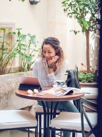 Téléchargez les photos : Souriant jeune femme assise dans un café en plein air et parlant sur tablette tout en ayant une conversation vidéo dans les médias sociaux dans la journée ensoleillée d'été - en image libre de droit