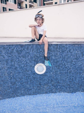 Photo for From below of full body of upset preteen boy in diving mask and flippers sitting on side on empty swimming pool and looking at camera unhappily - Royalty Free Image