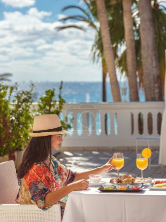 Téléchargez les photos : Vue latérale du voyageur féminin en chapeau assis à table avec petit déjeuner servi sur la terrasse de l'hôtel contre la mer pendant les vacances d'été - en image libre de droit