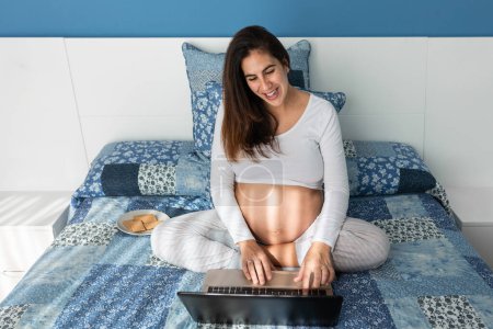 Photo for From above of smiling pregnant female speaking on video call on netbook while sitting on comfortable bed at home - Royalty Free Image