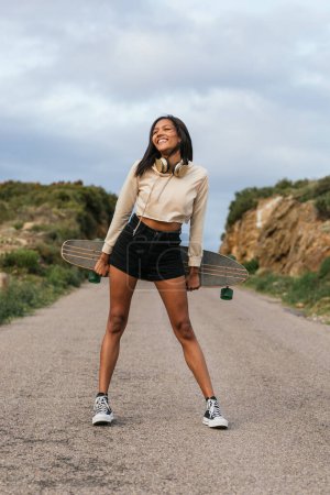 Photo for Full body of young smiling ethnic cheerful female standing with longboard and headphones on neck with eyes closed between hills under cloudy sky - Royalty Free Image