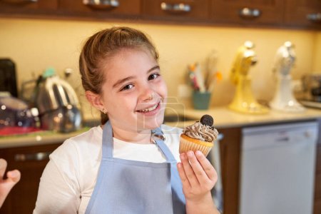 Téléchargez les photos : Enfant tressé positif avec le nez sale de crème portant tablier bleu et tenant gâteau maison savoureux décoré de pépites de chocolat et biscuit dans la cuisine et regardant la caméra - en image libre de droit