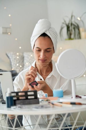 Photo for Adult focused woman with towel on head applying eyeshadow on hand while doing makeup and recording beauty blog on mobile phone at home in morning time - Royalty Free Image