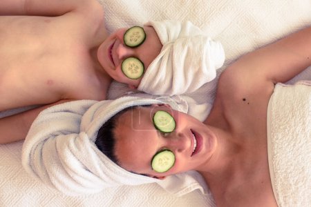 Photo for Top view of female and child resting on white blanket with towel on heads and cucumber slices on eyes - Royalty Free Image