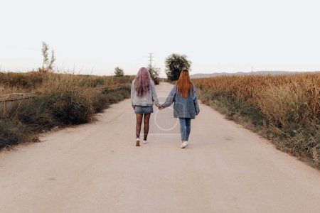 Photo for Back view of modern women in denim with long hair holding hands and walking on remote rural road - Royalty Free Image