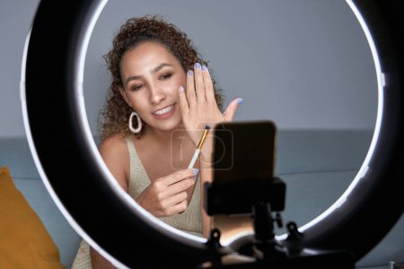Téléchargez les photos : Blogueuse de beauté féminine joyeuse montrant une brosse de maquillage professionnelle tout en enregistrant une vidéo de tutoriel sur smartphone et en utilisant une lampe annulaire - en image libre de droit