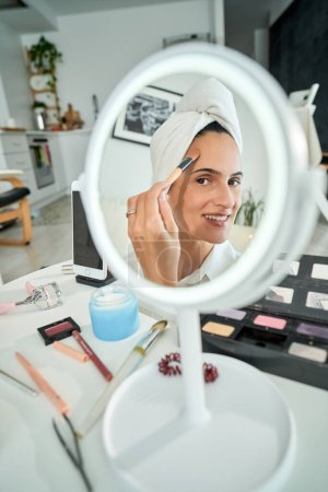 Téléchargez les photos : Angle élevé de réflexion dans le miroir de la femme souriante avec serviette sur la tête appliquer le fond de teint sur le visage avec une brosse tout en faisant le maquillage quotidien à la maison le matin - en image libre de droit
