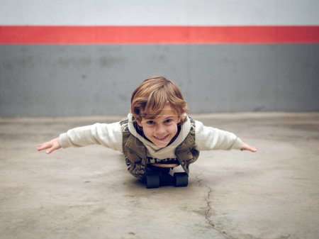 Foto de Adorable niño en sudadera con capucha acostado en monopatín con los brazos extendidos en vuelo en suelo de hormigón cerca de la pared en la calle mientras mira a la cámara - Imagen libre de derechos