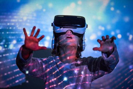 Photo for Amazed little child looking up with opened mouth and raising hands while exploring cyberspace in VR goggles standing near wall with colorful projection - Royalty Free Image