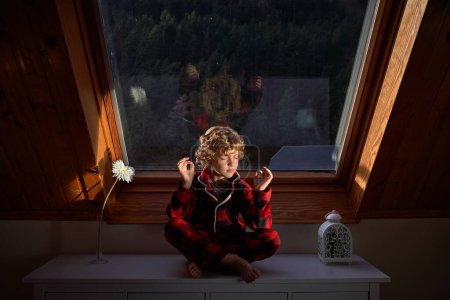 Photo for Full body of barefoot cute preteen boy meditating in Padmasana yoga pose while sitting with crossed legs on chest of drawers in evening - Royalty Free Image
