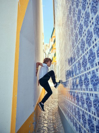 Photo for Side view full body of boy trying to scramble on decorative colorful wall of building located on narrow street in city - Royalty Free Image