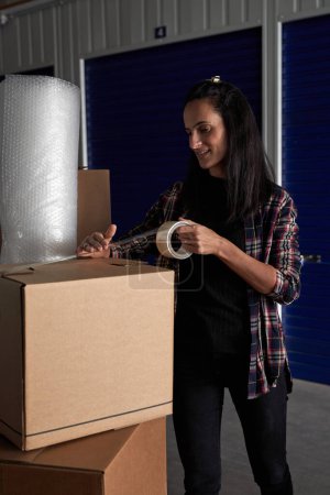 Photo for Content female in casual wear packing carton box with belongings using adhesive scotch tape neat self storage during moving day - Royalty Free Image