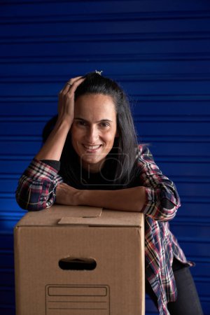 Téléchargez les photos : Femme joyeuse regardant la caméra avec le sourire tout en s'appuyant sur la boîte d'emballage en carton près des portes fermées de self-stockage pendant la journée de déplacement - en image libre de droit