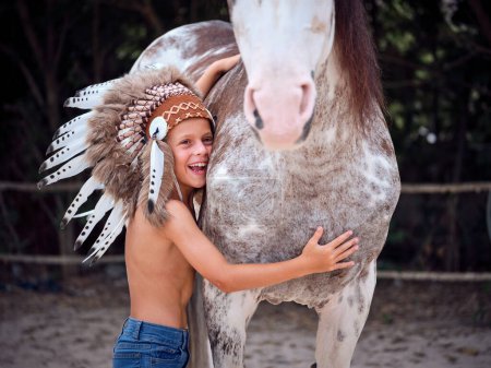 Foto de Vista lateral de niño alegre preescolar en jeans de pie en suelo arenoso y abrazando alegremente caballo blanco de pura raza - Imagen libre de derechos