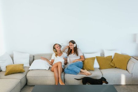 Photo for Positive female friends with smartphone singing while listening to music in wireless headphones on comfortable couch in light living room - Royalty Free Image