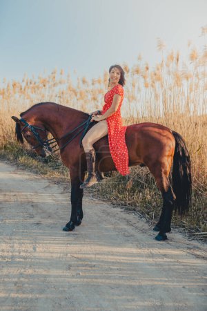 Foto de Joven hembra en vestido rojo sonriendo y mirando a la cámara mientras está sentado en el caballo en el camino del campo cerca del campo - Imagen libre de derechos