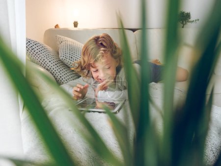 Foto de A través de hojas verdes de plantas vista de niño preadolescente sonriente acostado en el sofá y tableta de navegación - Imagen libre de derechos