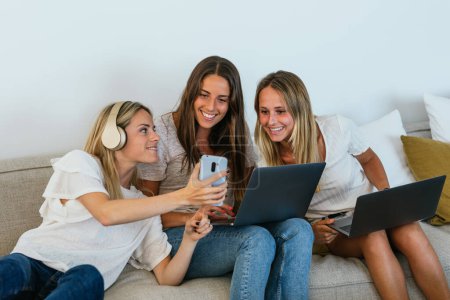 Foto de Mujer en auriculares que muestran video en el teléfono celular a alegres amigas con computadoras portátiles mientras están sentadas en el sofá en la sala de estar - Imagen libre de derechos