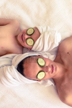 Photo for From above of relaxed female and child lying on bed with towel on heads and cucumber slices covering eyes - Royalty Free Image