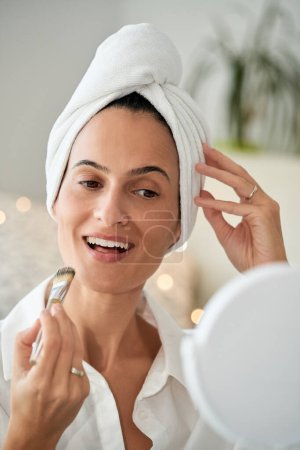 Téléchargez les photos : Délicieux adulte souriant femelle avec serviette sur la tête appliquer le fond de teint à l'aide d'une brosse gagner devant le miroir tout en touchant la tête à la maison le matin - en image libre de droit