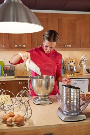 Téléchargez les photos : Chef pâtissier femelle regardant vers le bas tout en remplissant bol avec de la crème à l'aide de spatule près de la table dans la cuisine - en image libre de droit