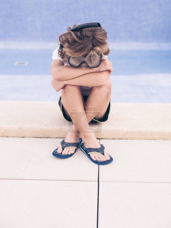 Foto de Cuerpo completo molesto chico en el buceo googles y zapatillas abrazando las rodillas mientras se sienta contra la piscina - Imagen libre de derechos