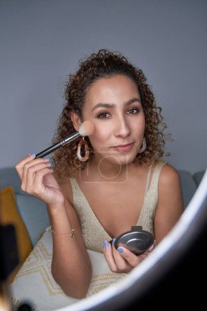 Photo for Charming curly haired makeup master woman showing shading on glow highlighter powder on cheek with professional brush while recording video - Royalty Free Image