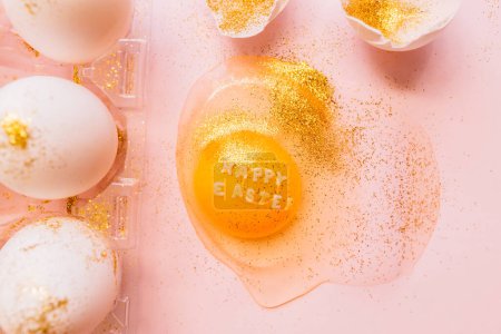 Photo for Top view of broken glittered egg with text Happy Easter and raw eggs in plastic box on pink background - Royalty Free Image